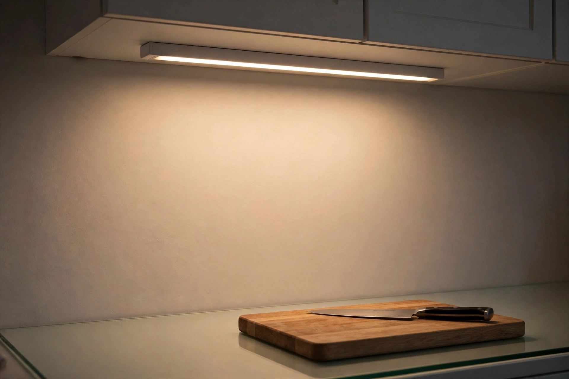 a slim battery light bar glowing warm white under a kitchen cabinet, lighting a wooden chopping board and knife on the counter below
